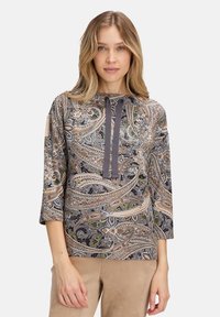 Kobieta w bluzce w wzór paisley z rękawami do trzy czwarte i ciemnymi sznurkami, w połączeniu z beżowymi spodniami, stojąca na tle białym.