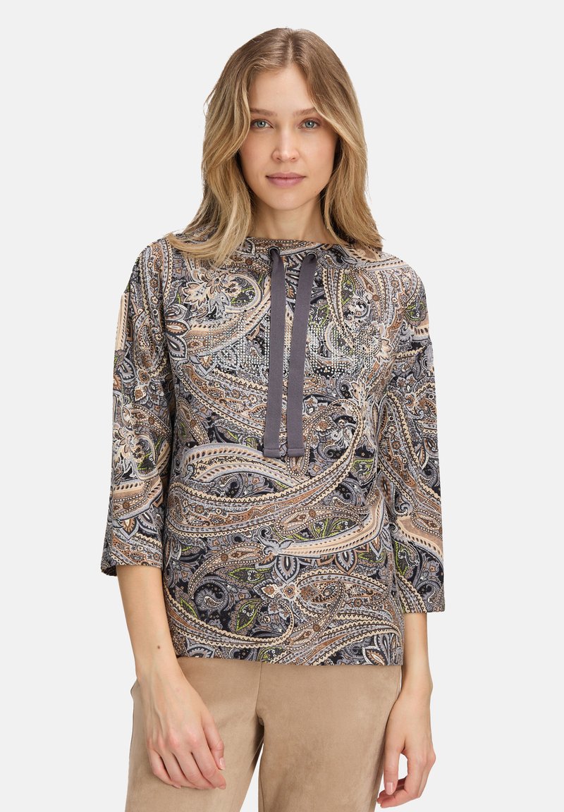 Kobieta w bluzce w wzór paisley z rękawami do trzy czwarte i ciemnymi sznurkami, w połączeniu z beżowymi spodniami, stojąca na tle białym.