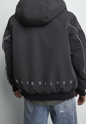 Personne portant une veste à capuche Quiksilver noire avec des passepoils réfléchissants sur les manches et un jean bleu clair déchiré, vue de dos.