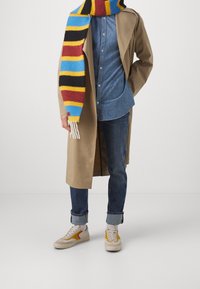 Beige trench coat, blå jeansskjorta, mörka jeans, flerfärgad randig scarf och beige sneakers med röda och gula accenter.