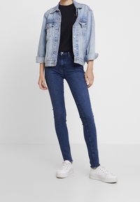 Chaqueta de mezclilla en azul claro, camiseta negra ajustada, vaqueros skinny de tiro alto en azul oscuro y zapatillas blancas con suela texturizada.