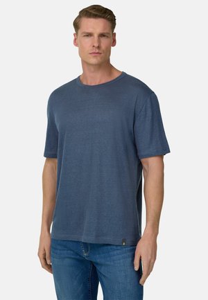 Homme aux cheveux courts châtain clair portant un T-shirt bleu uni à manches courtes et un jean bleu, debout devant un fond blanc.