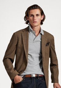 Blazer de espiga marrón sobre una camiseta polo gris, con un pañuelo negro en el bolsillo, jeans oscuros y un cinturón marrón con hebilla plateada.