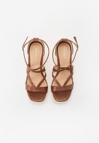 Sandales en cuir marron avec plusieurs brides croisées, un bout carré et un petit talon carré. Elles sont dotées d'une sangle à la cheville avec une boucle.