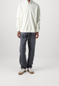 Pullover bianco in pile con design a zip a un quarto, maniche lunghe, logo sul petto, abbinato a jeans dritti grigio scuro e sneaker.