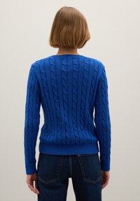 Blauwe kabelgebreide trui met geribde zoom en manchetten, voorzien van een gestructureerd patroon en een ronde halslijn. Draagt bij donkere spijkerbroeken.