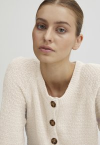 Világos bézs kötött kardigán, texturált felülettel, kerek nyakkivágással és öt fa gombbal elöl.
