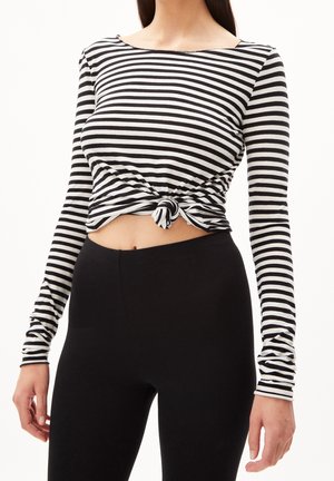 Mujer con una camisa de manga larga a rayas blancas y negras anudada en la cintura combinada con leggings negros de talle alto sobre un fondo blanco.