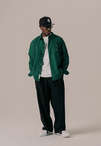 Zöld ing fehér póló felett, fekete bő szárú nadrággal és fehér tornacipővel párosítva. Kiegészítők: fekete sapka és szemüveg.