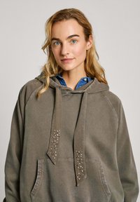 Sudadera gris con bolsillo tipo canguro y cordones decorados con remaches plateados. Tela suave y texturizada con mangas raglán.