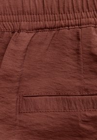 Roodbruine stof met een zachte textuur, voorzien van een elastische tailleband en een enkele gestikte zak. Het ontwerp is eenvoudig en functioneel.