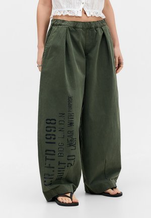 Løse mørkegrønne bukser med sort tekst på det ene ben, båret med sorte sandaler og en hvid cropped top med blondedetalje.