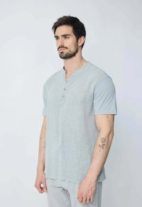 Chemise bleu clair à manches courtes avec une surface texturée, dotée d'un col henley et de trois boutons en bois. Portée avec un pantalon ample.