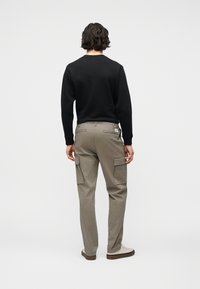 Άτομο με κοντά σκούρα μαλλιά, που φοράει μαύρο μακρυμάνικο πουκάμισο, μπεζ παντελόνι τύπου cargo και ανοιχτόχρωμα παπούτσια, στέκεται με την πλάτη προς το μέρος μας σε απλό φόντο.