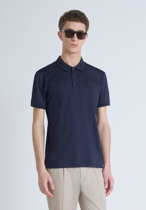 Camisa polo azul marino con un patrón de Chevron texturizado, tapeta de tres botones y mangas cortas. Combinada con pantalones beige claros.