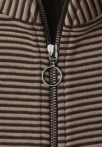 Chaqueta de punto con cremallera de rayas en beige y marrón con una cremallera plateada prominente y un tirador circular. La textura parece suave con detalles acanalados.