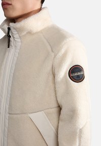 Cream-colored fleece jacket with a high collar, side panel accents, and a circular logo patch on the upper arm. Soft, textured fabric.