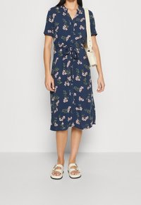 Navy blue short-sleeve dress with a floral pattern, belted at the waist. Paired with white slide sandals featuring decorative hardware.