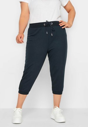 Persoon draagt zwarte cropped sweatpants met taillekoord en witte sneakers, staand met één hand op de heup.