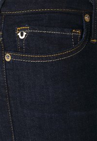 Jean de mezclilla azul oscuro con costuras doradas en contraste y un pequeño logotipo blanco en el bolsillo delantero. La tela tiene un acabado suave y texturizado.