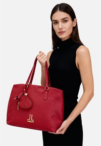 Borsa tote in pelle rossa con doppi manici, dotata di dettagli in oro. Include un piccolo charm a forma di cuore con motivo trapuntato e marchio.