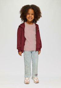 Lachend kind met krullend haar draagt een bordeauxjack over een roze shirt, lichtblauwe spijkerbroek en roze-witte sneakers, staande tegen een effen achtergrond.