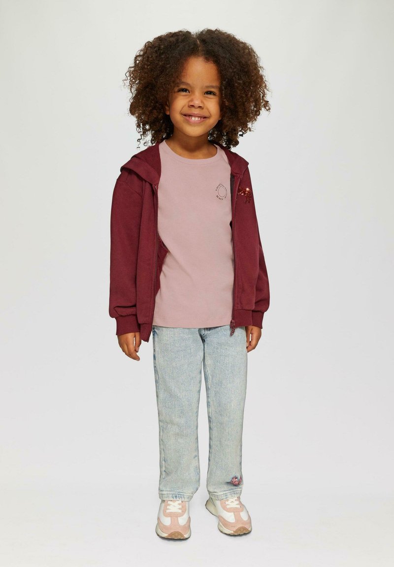 Lachend kind met krullend haar draagt een bordeauxjack over een roze shirt, lichtblauwe spijkerbroek en roze-witte sneakers, staande tegen een effen achtergrond.