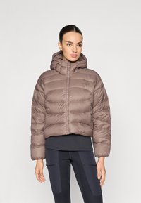 Brun puffjacka med huva, prydd med ett kviltat mönster, framdragkedja och elastiska ärmslut. Bärs över en mörk topp och kombineras med svarta leggings.