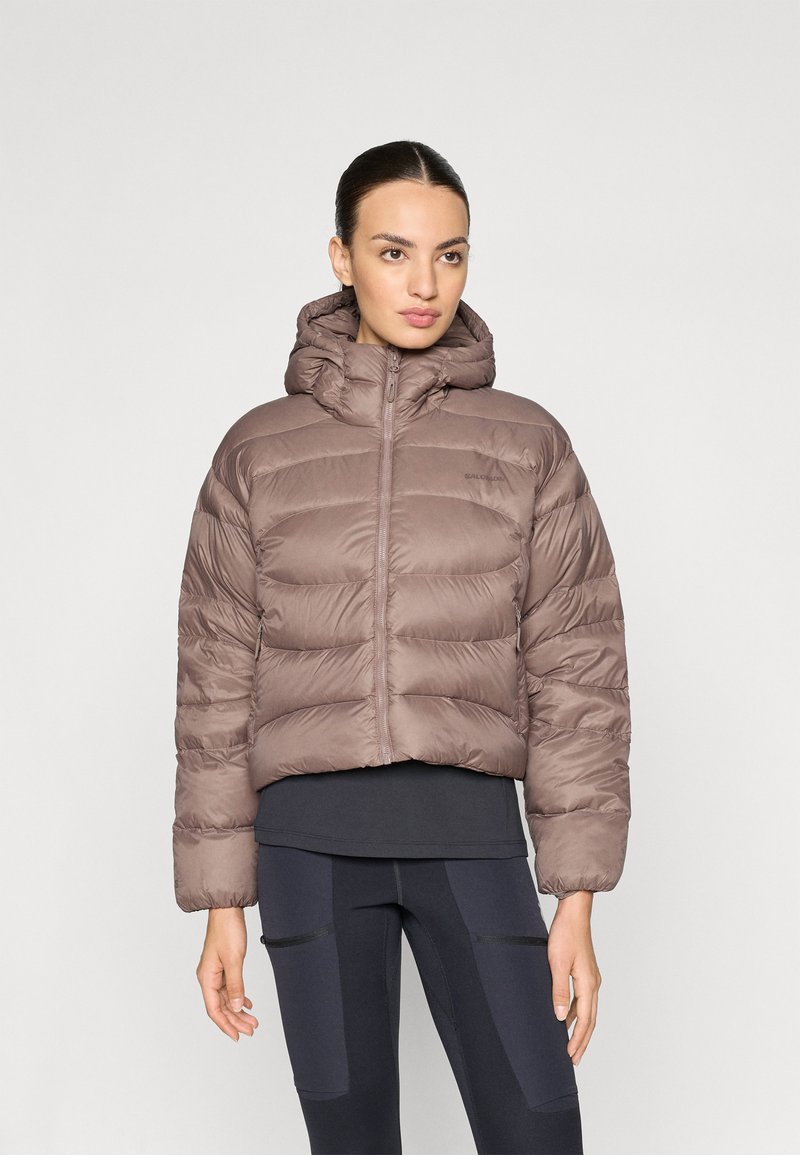 Brun puffjacka med huva, prydd med ett kviltat mönster, framdragkedja och elastiska ärmslut. Bärs över en mörk topp och kombineras med svarta leggings.