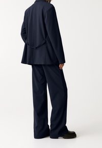 Personne debout de profil portant un costume bleu marine foncé à fines rayures avec un pantalon à jambes larges et des chaussures noires épaisses sur un fond uni.