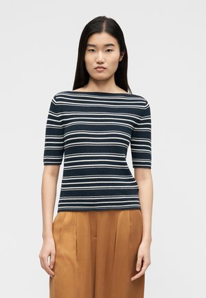 Femeie cu păr lung și închis la culoare, purtând un top cu dungi albastru închis și alb, cu decolteu bateau și mâneci până la cot, și pantaloni maro plisati.