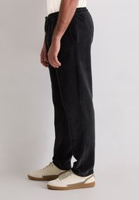 Ein Mann steht in schwarzen Cordhosen mit Taschen und off-weißen Sneakern mit braunen Sohlen vor einem schlichten, hellen Hintergrund.