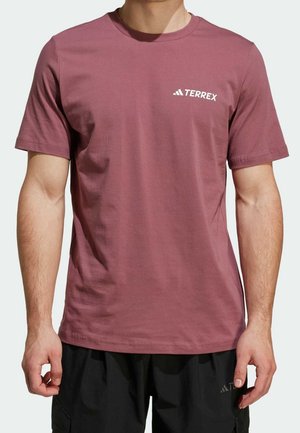 Homme portant un t-shirt Terrex rose poussiére à manches courtes et un short noir sur un fond clair uni.