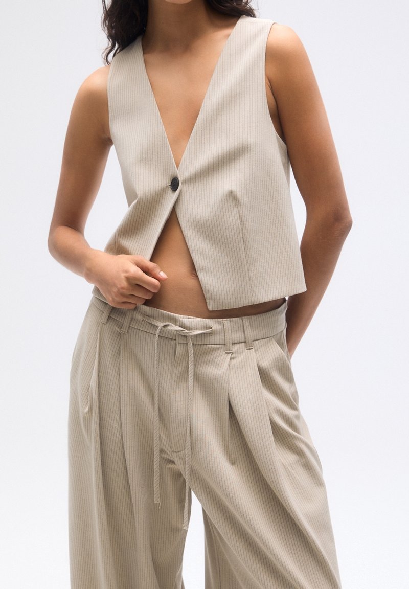 Gilet cropped à rayures beiges avec un décolleté en V, doté d'une fermeture à bouton. Assorti à un pantalon ample à rayures assorties avec cordon de serrage.