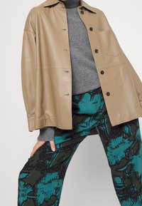 Beige leren jas met een relaxte pasvorm, grijze gebreide trui en een broek met een patroon in teal en donkergroene bloemmotieven.