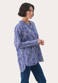 Niebieska i biała bluzka w print, z kołnierzykiem stojącym, długimi rękawami i luźnym krojem. Posiada wzór paisley i zaokrąglony dół.