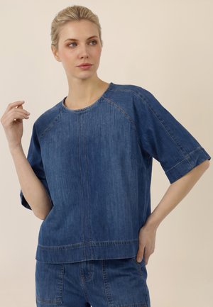 Frau mit blondem Haar, die ein locker sitzendes blaues Denim-Oberteil und passende Denim-Hosen trägt und mit einer Hand nahe ihrem Gesicht posiert.