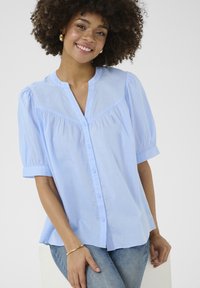 Camicia blusa azzurro chiaro con maniche corte a sbuffo, collo arrotondato e dettagli arricciati lungo il busto. Tessuto morbido e leggero.