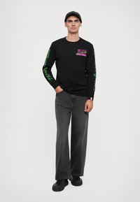 Camisola preta de manga longa com detalhes gráficos em roxo e verde no peito e mangas, combinada com jeans largos cinza e sapatos pretos.