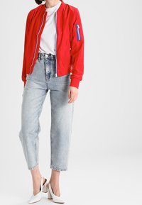 Veste bombardier rouge avec une fermeture éclair bicolore, t-shirt blanc, jean taille haute bleu clair, et mules blanches à talons.