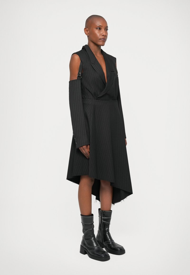 Schwarzes Kleid mit Nadelstreifen, asymmetrischem Saum, tiefem V-Ausschnitt und offenem Schulterbereich. Kombiniert mit knielangen, schwarzen, glänzenden Stiefeln.