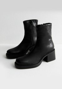 Botas de tobillo de cuero negro con tacón cuadrado, presentan una cremallera lateral, textura suave y detalles de costura a lo largo de la suela.