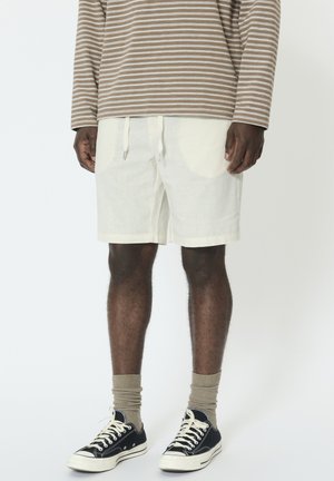 Muž nosiaci béžovú a bielo pruhovanú košeľu s dlhým rukávom, krémové šortky so sťahovacou šnúrkou, béžové ponožky a čierne nízke tenisky, stojí nehybne.