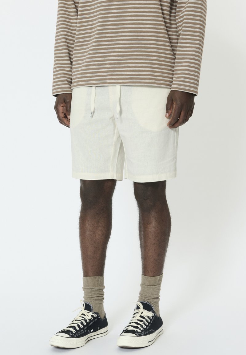 Homme portant une chemise à manches longues rayée beige et blanche, un short écru avec cordon de serrage, des chaussettes beiges et des baskets basses noires, debout immobile.
