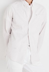 Jasnoróżowa koszula z białymi paskami, zapinana na guziki, z długimi rękawami, spiczastym kołnierzykiem i jedną kieszonką na piersi, zestawiona z białymi spodniami.