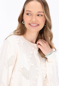 Cardigan a maglia crema con motivi a cuore testurizzati, accenti a pompon e bottoni a forma di cuore. Modella che indossa orecchini e bracciali di perline.