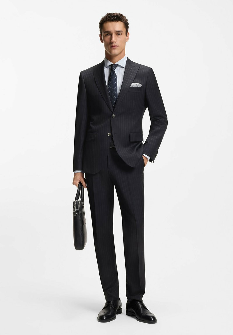 Garnitur w granatowy prążek o kroju dopasowanym, zestawiony z jasnoniebieską koszulą w paski i krawatem w kropki. Czarne skórzane buty i czarna teczka.
