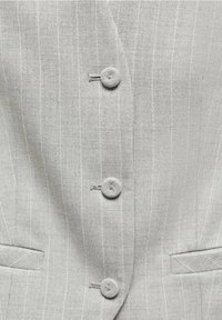 Gros plan d'un blazer gris clair à fines rayures montrant trois boutons recouverts de tissu et deux poches fentes horizontales.