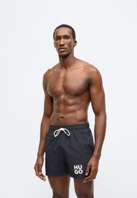 Tamsiai mėlyni maudymosi šortai su raišteliu juosmenyje, ant kairės kojos baltos spalvos „HUGO“ užrašas, pagaminti iš lengvos, lygaus audinio tekstūros.