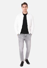 Giacca bianca trapuntata sopra un maglione nero, abbinata a jeans grigio chiaro e sneakers nere. La giacca ha una chiusura con zip e un colletto alto.